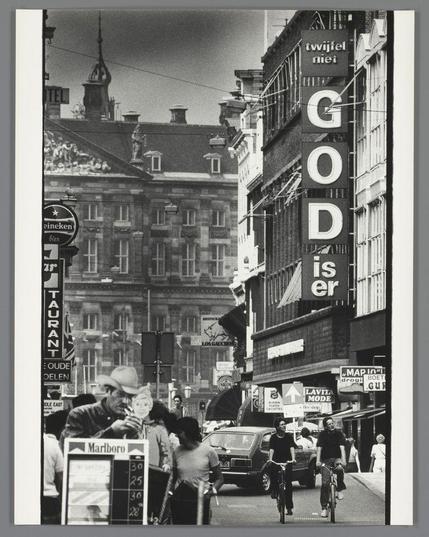 The image depicts a bustling urban street scene, likely taken in Amsterdam during the early 1980s. The photograph captures pedestrians and cyclists going about their daily activities on what appears to be Damstraat, characterized by its historic buildings with ornate facades.

On either side of the street are various shops and establishments, including "GOD is er" (God Is Here) in prominent lettering above one storefront, which could suggest a religious or spiritual message. The wordmark also includes the phrase 'twijfel niet', translating to 'do not doubt' from Dutch, indicating an encouragement for faith.

Pedestrians are dressed casually and some engage with each other while others focus on their surroundings. One man in the foreground is seen smoking a cigarette and another cyclist appears to be riding past him, alluding to contemporary urban life. The presence of a "Marlboro" billboard indicates commercial activity and advertising prevalent at that time.

The black-and-white nature of the photo adds a timeless quality, emphasizing textures and contrasts rather than colors. This visual style is reminiscent of street photography popularized by photographers like Gjon Mili and Lee Friedlander during their respective periods.

Overall, this image provides a glimpse into urban life in Amsterdam from around 1980-1990, showcasing the blend of historical architecture with modern commercial activities against an everyday backdrop.