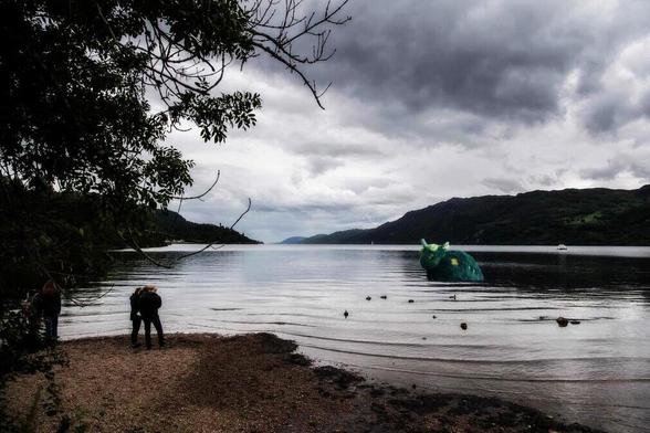 A photograph of Loch Ness with moody clouds, and a plush creature in the water, while onlookers look on at the shoreline.
