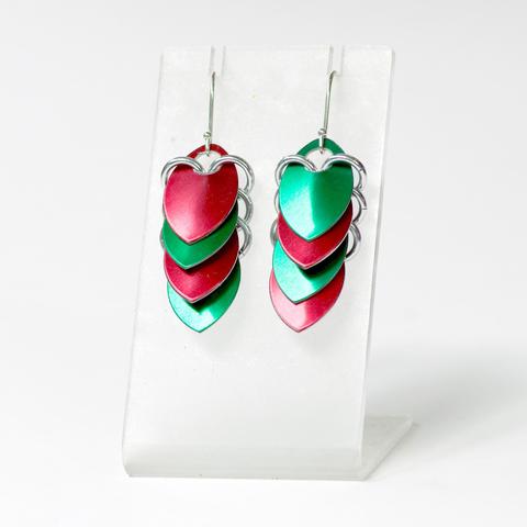 A pair of scalemail earrings on a white display stand; each earring is red and green, but they alternate stripes so one has red at the top and one has green at the top.