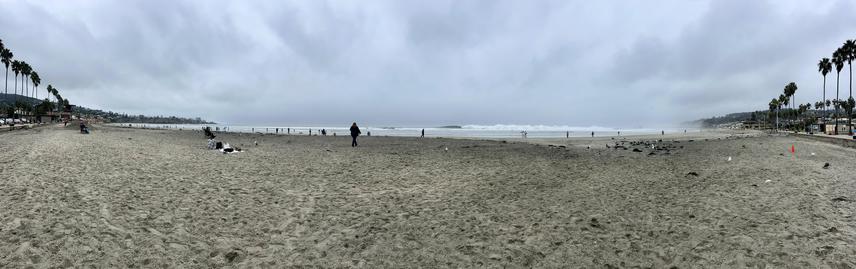 Panorama of the beach with overcast skies, La Jolla to the pier in the far distance. Scattered people on the beach and breaking waves.
