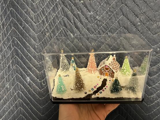 A white person’s hand holding up a 6.5” long miniature display case diorama of a gingerbread house in a forest of snowy multicolored pine trees.