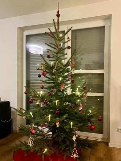 Ein geschmückter Weihnachtsbaum mit roten Ornamenten, Lichtern und verschiedenen festlichen Dekorationen. Der Baum steht in einem gut beleuchteten Raum mit großen Fenstern. Eine rote Baumschürze bedeckt den Sockel.