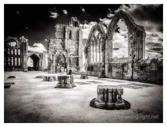 A toned black and white infrared photo of the interior of a ruined cathedral. the north and west walls and their window frames can be seen, and the stumps of pillars.
