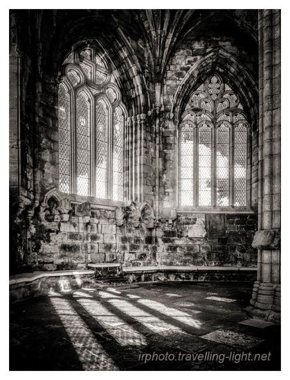 A toned black and white infrared photo of the interior of the Chapter House of a ruined cathedral. There are tall windows with restored glazing.