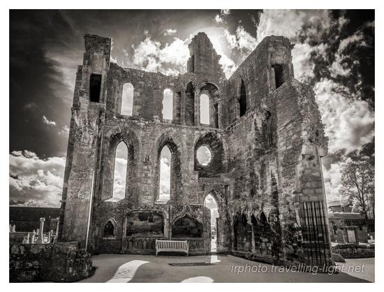 A toned black and white infrared photo of the interior of a ruined cathedral, lookig into the sun, which is hidden behind part of a wall.