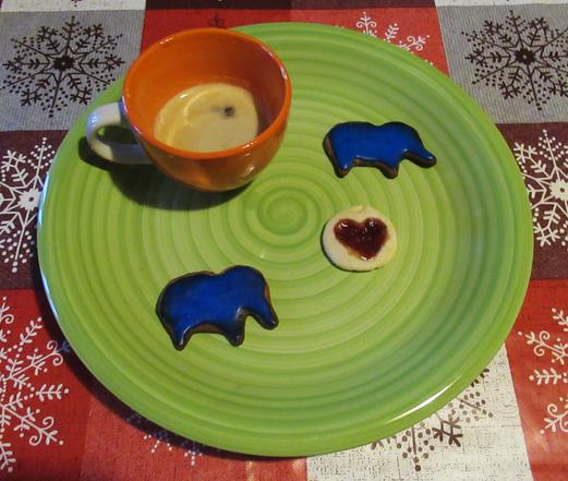 Ein großer grüner Teller. Darauf zwei Kekse in Form eines Rüsseltieres mit blauem Zuckerguss und ein roter Keks mit einem roten Herz aus Marmelade. Eine Tasse mit Espresso.