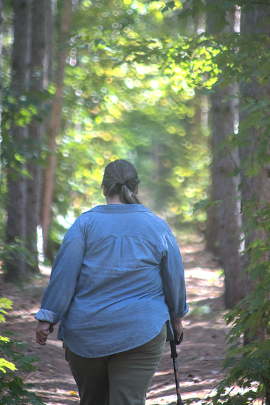 Julie walks along a wooded trail at Aspen Park in Gaylord, MI, with tall trees lining the path. She is seen from behind, wearing a light denim shirt and khaki pants, holding a walking stick in one hand. The sunlight filters through the leaves, creating a soft, dappled glow and a peaceful atmosphere. The trail is surrounded by vibrant green foliage.