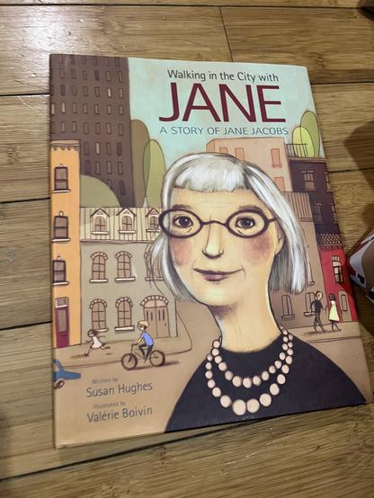 Close up color photo of a children’s book titled “Walking in the City with Jane” with an illustrated image of an older light skinned woman with short gray hair and glasses with an urban street background