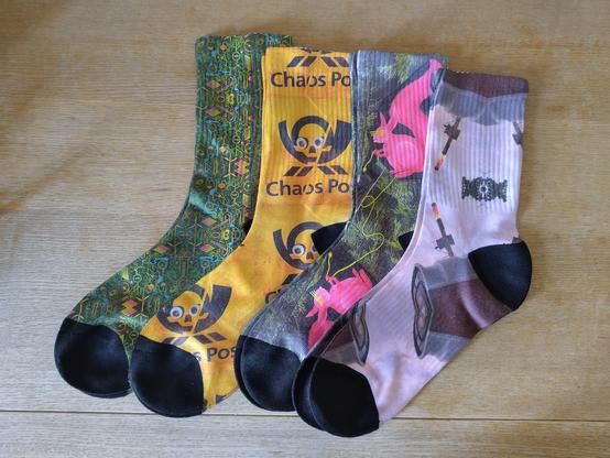 Four socks in various chaos adjacent designs - EMF 2024 solarpunk design, chaos post logo with googly eyes, the dreaded cybersquirrel nibbling on some tasty glasfaser and a permuted datenklo.