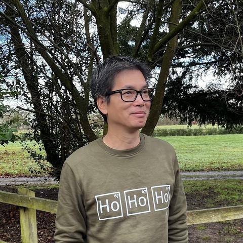 A person wearing a green sweatshirt with a humorous periodic table design featuring holmium that spells out "Ho Ho Ho" stands outdoors near a tree, with a grassy area in the background.