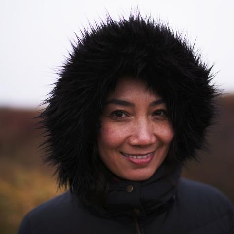 Portrait of a lady, looking in to the lens and sporting a cheeky smile. Her hood seems a bit spiky, the background is blurred with yellow, brown and grey tones.
