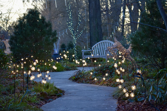 A winding stone walkway bordered by round white Christmas bulbs leads to a pale gray bench. Tiny white lights decorate the surrounding evergreen trees and bushes.