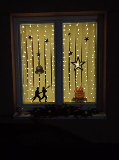 Ein beleuchtetes Fenster. Auf dem Fenster sind zwei Feuerwehrleute mit Strahlrohr, ein klassischer Feuerwehrhelm "Wiener Form", mehrere Sterne, die Zahl 21 und ein kleines Feuer aufgeklebt. 

Auf dem Fensterbrett davor liegen Tannenzweige und Christbaumkugeln