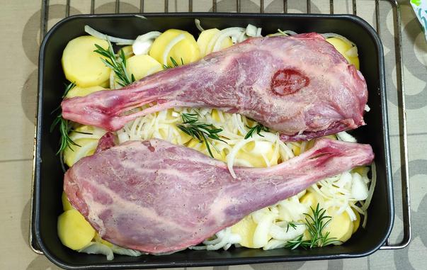 Safata de forn amb dues cuixes de cabrit sobre un llit de patata i ceba, abans de rostir-les.