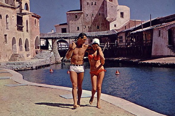 1965 Vintage Beach Romance
Description: This image depicts a couple dressed in swimwear from an earlier era, walking alongside what appears to be a Mediterranean-style village with water and sailboats. The man is shirtless wearing white swimming trunks while the woman sports a red bikini top and orange bottoms.