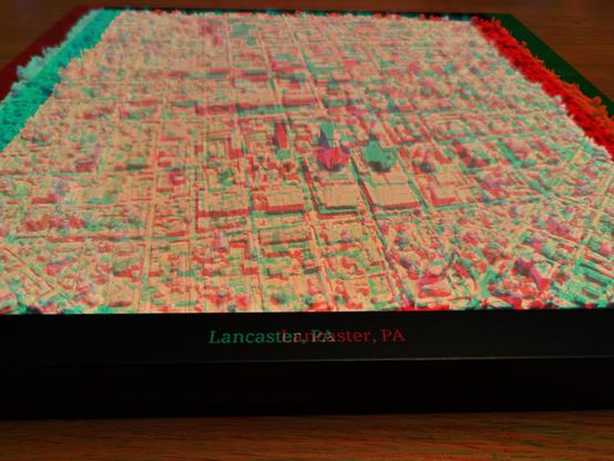 Closeup of a white, 3D printed relief of Lancaster City, Pennsylvania in a black frame, with the legend “Lancaster, PA” printed in silver lettering on the frame. Presented as a Red/Blue Anaglyph stereoscopic image.