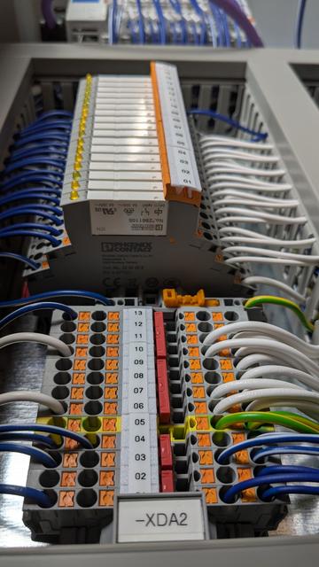 Part of a control panel with terminal blocks at the bottom and DIN rail relays above.  Lots of dark blue and white wires terminate at the devices.  Wire ducts run all around them.  All terminal blocks and relay modules are labelled.