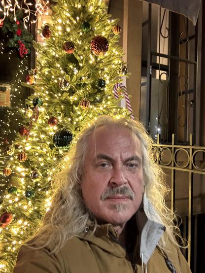 Man in a selfie, long white hair, salt and pepper mustache/ jazz patch, with a grey respirator mask hanging from one ear. He’s standing in Manhattan in front of a restaurant/ hookah place who has a huge decorated tree with color changing LED lights. In the photo the lights are all warm white.