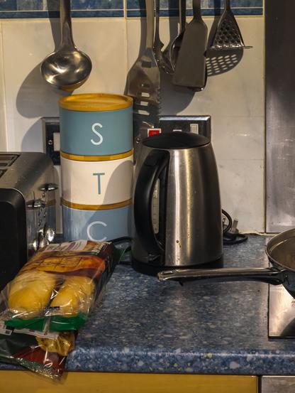 In a kitchen, three round jars are stacked on top of each other, with the letters S, T and C on them from top to bottom. There's other things asking it – kettle to one side, toaster to the other, baguettes behind it, and spatulas above.