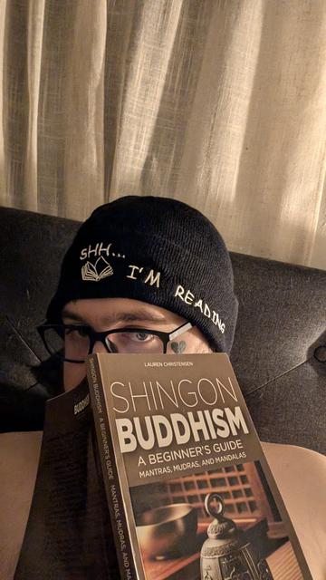 A person is partially visible, wearing glasses and a black beanie that says, "Shh... I'm reading," with an open book graphic embroidered on it. They are holding up a book titled Shingon Buddhism: A Beginner's Guide - Mantras, Mudras, and Mandalas by Lauren Christensen. The setting appears to be a dimly lit, cozy environment with beige curtains in the background.