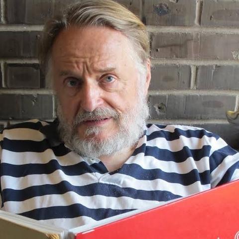 This image shows John Horton Conway, a renowned mathematician, seated and holding an open book with a red cover. He has a thoughtful expression, slightly raising his eyebrows. Conway has a full white beard and wears a navy blue and white striped shirt. The background features a brick wall, adding a casual, home-like setting to the scene. Conway is best known for his contributions to mathematics, including the creation of the "Game of Life," a cellular automaton that explores complex systems.
(description created with chatGPT but reviewed by myself).