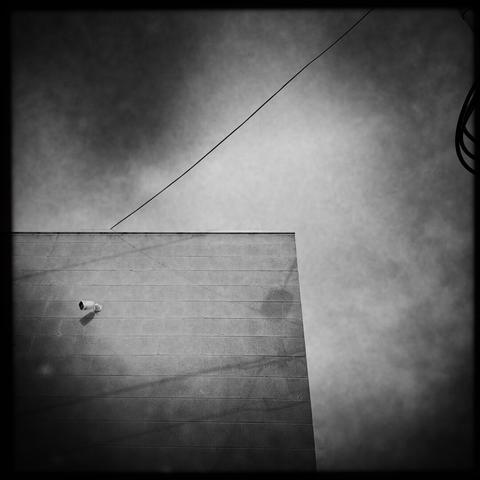 A murky black & white photo of a building with a grey wall, containing a security camera looking off to the left. The shadow of a few wires plays across the wall, and one of the wires is visible tracing a path from mid-left up to top-right.