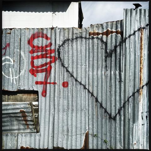 A silver corrugated fence, with the outline of a heart in black over to the right, some graffiti in red on the left (maybe saying C37), and at the bottom left a missing panel. The shadow of a roof is above, and a cloudy sky.