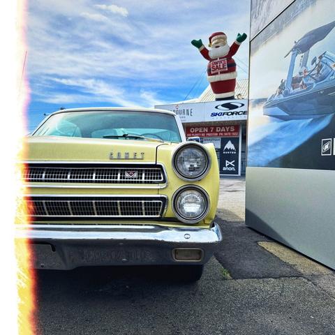 The front of a yellow 1960s car, one headlight above the other. It says “Comet” and a badge underneath suggests it’s a V8. In the distance beyond, an inflatable Santa has his arms up, celebrating in front of a cloudy streaky sky.