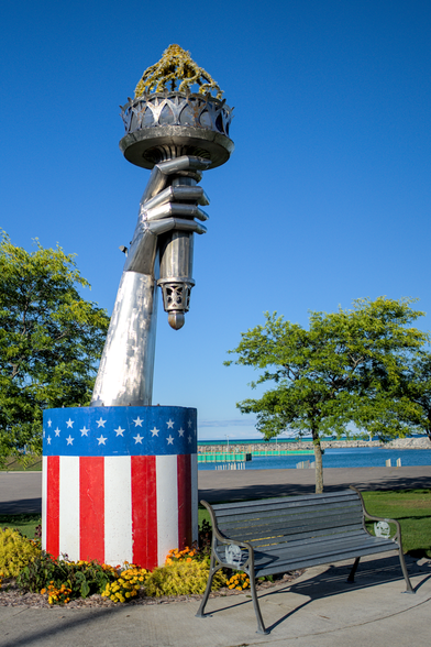 A large sculpture in Rogers City, MI, resembling an outstretched hand holding a torch, styled to evoke the Statue of Liberty. The torch is adorned with metallic elements and a cascading floral arrangement at the top. The base is painted with red, white, and blue stripes and stars, symbolizing the American flag. Surrounding the sculpture, vibrant yellow flowers and a neatly maintained landscape add color to the scene. A black bench sits nearby, with a clear view of Lake Huron and a bright blue sky in the background.