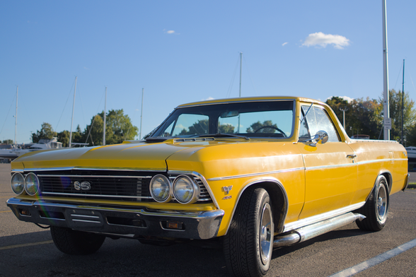 A classic yellow Chevrolet El Camino SS, with sleek chrome accents and SS badging on the grille, is parked in Rogers City, MI. The car's polished exterior shines in the sunlight, showcasing its vintage charm. In the background, sailboats and masts are visible against a clear blue sky, with trees adding to the scenic harbor setting.