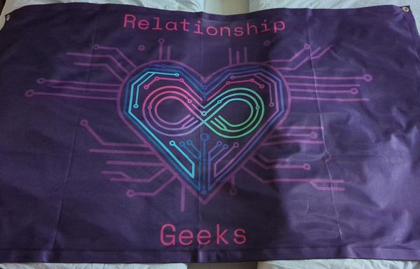 A flag with the Relationship Geeks logo, lying on a bed. The flag shows a heart symbol embedded in computer circuit lines. Within the heart, there is an infinity symbol created out of more circuit lines. The logo is colorful on a dark purple background and gives of a happy, geeky vibe.