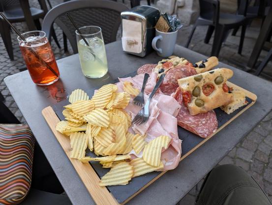 A table outside a café upon which is a mug with sugar packets, a napkin dispenser, a Campari spritz, a limoncello spritz, and a huge platter sufficient for four people with potato chips, pistachio mortadella, porchetta, salami, and olive and cherry tomato focaccia.