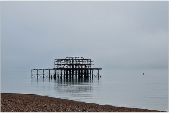 Skeleton of a building in a calm sea, the remains of a pier that burned down, with a narrow slice of a pebble beach in the foreground.