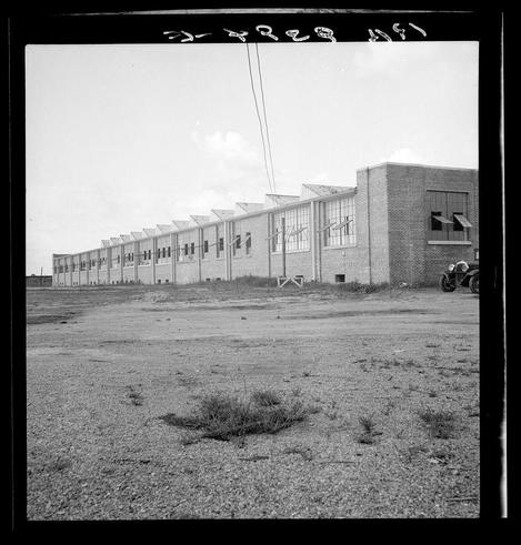 The image is a black and white photograph depicting what appears to be an industrial facility or factory. The structure has multiple rows of windows along its length, with the middle section appearing taller than the wings on either side.

In front of this building lies an open area that looks somewhat barren, with sparse vegetation visible in patches across the ground. To the right foreground of the image, a motorcycle is parked near what could be interpreted as part of another structure or barrier made from metal and concrete elements.

The sky above appears clear without any noticeable cloud cover. The overall condition suggests it might be an old facility given its architecture and lack of maintenance in certain areas such as the ground's appearance.