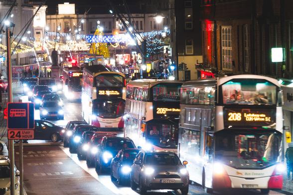 Night street scene in Cork city during Christmas season showing heavy traffic with buses (routes 205, 207, 208, 212) and cars, illuminated by festive overhead lights and decorations