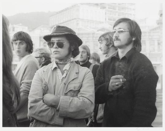 The black and white photograph captures a group of individuals in what appears to be an outdoor urban setting, likely during the late 1960s or early '70s based on their attire. In focus are four men with serious expressions standing side by side; they seem contemplative or concerned as if awaiting something significant.

The man at center-left wears sunglasses and a wide-brimmed hat, exuding an air of nonchalance or perhaps defiance, contrasting the more somber demeanor around him. His relaxed posture contrasts sharply with his companions' attentive stances. The two men to his right are dressed in dark clothing; one is wearing glasses and has a mustache while clutching something small between his fingers.

The background suggests an urban environment bustling with activity or possibly protest, as inferred from the individuals present but not focused on by the photographer's attention. A woman stands at far left edge of frame partially cropped out—her presence adds to atmosphere yet remains anonymous due to her peripheral inclusion within this moment captured in time.

This image is notable for its candid portrayal of an apparent social or political gathering, offering viewers a window into historical activism and public expressions during that era.