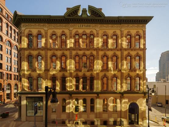 The Ledyard building, a historic commercial structure, is bathed in the warm light of the late afternoon sun. Reflections of sunlight from the windows of a neighboring building cast intricate patterns of light and shadow across the numerous arched windows, creating a dynamic interplay of light and dark on the light beige brick facade. The decorative elements, including the ornate window frames and the sculpted details above the windows, add to the building's architectural character.