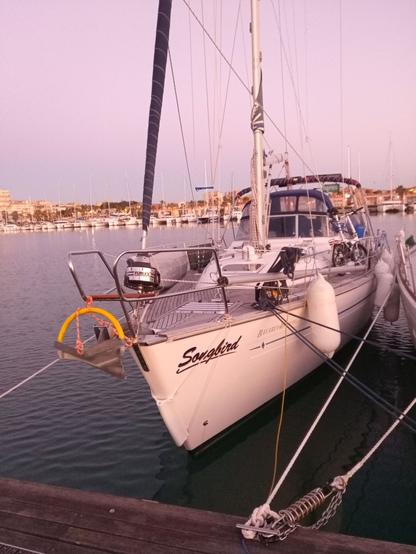 A 40 ft sailboat in its winter berth in Sicily. Things are lashed down for bad weather, but the water is calm. There's a folding bicycle on deck. Rows of boats in the distance, behind a town on a hillside.