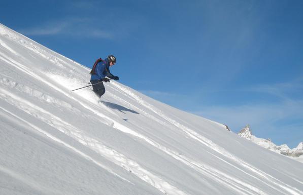 Photo of a telemark skier skiing down a slope of fresh snow with blue sky and the peaks of a few mountains in the background.