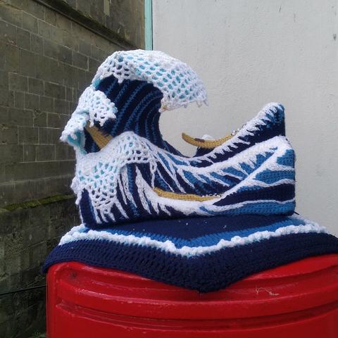 A representation of Hokusai's The Great Wave rendered in knitting. It's a work of genius (the knitting). Prussian blue and white for the wave, and brown/amber for the boats.
It sits on top of a postbox which is Royal Mail red. Immediately behind is a white painted rendered wall, and to the left background a bit further away is the side of a greenish stone building.