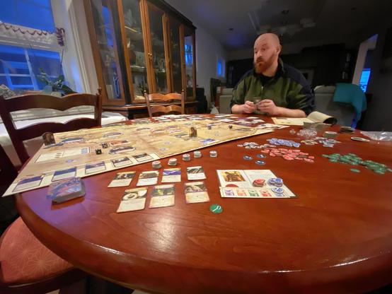 A man sits at a wooden table with a board game spread out in front of him. The table features various game cards, tokens, and dice, with a scenic game board visible.