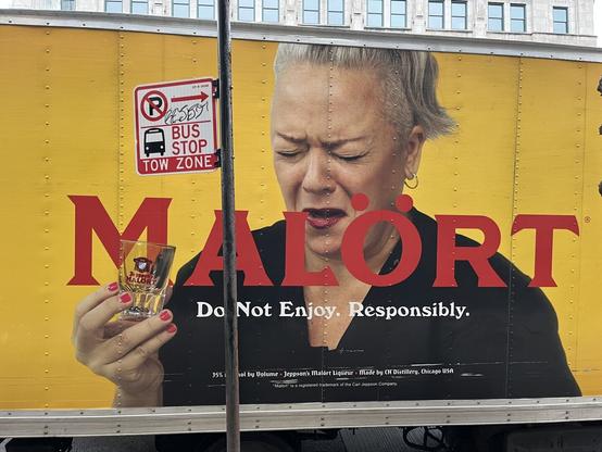 An advertisement on the side of a vehicle. It has a woman with a shot glass on her hand with a grimace on her face. Over her image is the word "MALÖRT" in red letters. Underneath is the phrase "Do Not Enjoy. Responsibly" in white letters. Everything is on a mustard yellow background.