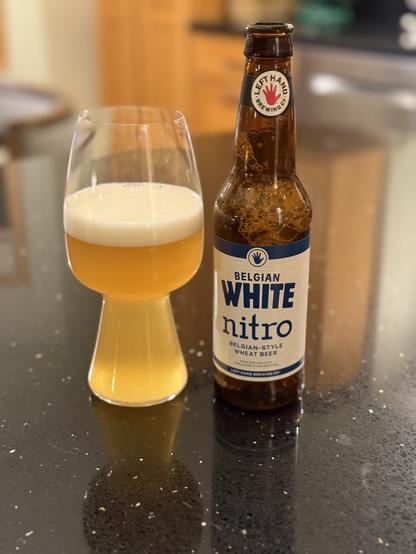 A stout glass of golden hazy beer. The brown bottle has a white label with dark blue lettering and the iconic red Left Hand logo on the neck