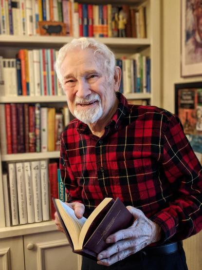 Robert Wolke, white haired and goatee, in a red checked flannel shirt, smiling at the camera, with a bookshelf in the background