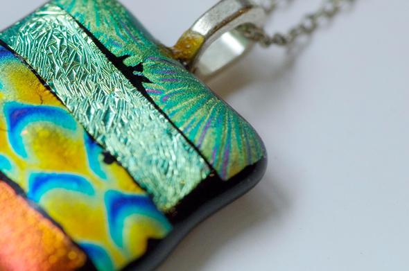 A squareish pendant, angled; it consists of four textured stripes in shades of iridescent green, gold, blue and orange. Set against a white background.