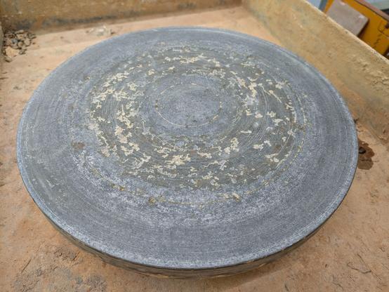 A photo of a circular pottery throwing wheel plate. It is a grey colour. The surface is pitted with craters, especially in a ring about 1/3 out from the centre.