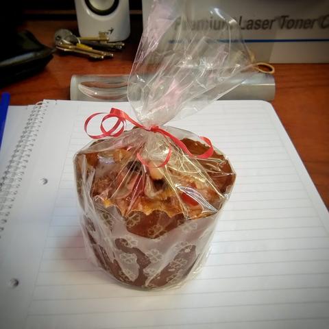Mini pan dulce envuelto en celofán transparente, con un pequeño lazo rojo, sobre un cuaderno abierto en el escritorio.