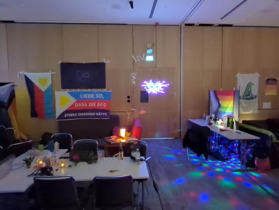A photo of the relationship geeks assembly space. Taken so that no people are visible.

On the right there are two desks: one with tea and biscuits, and one with the question box, waiting for your thoughts. Underneath the latter there is a light throwing colourful reflections dancing over the floor.

On the left there is in the foreground a table and chair setup, serious work is done here. In the background, there is the comfy RG Lounge area with couch and mattresses, in which I'm typing this.

On the wall in the background, there are a couple of flags, the progress pride flag, the Relationship Geeks flag, a polyamory flag and a very special custom polyflag with writing on it, spelling: "Love in a way that the afd would oppose.