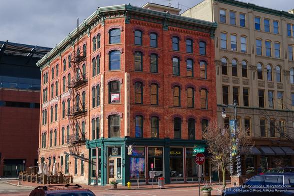 A photograph of a street scene shows a corner of a historic red brick building, likely a commercial or residential structure, with various businesses. The building, with its ornate brickwork and arched windows, is five stories tall. A sign in a window advertises available office space for lease. A restaurant, "Lounge," is visible on the corner, with its neon sign and outdoor seating area. Other businesses are also visible, judging by the signage and awnings. A stop sign is present at the street intersection. Cars are parked on the street, and a single pedestrian is visible on the sidewalk.