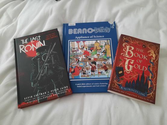 Three books on a white bed. The Last Ronan by Kevin Eastman and Peter Laird (Ninja Turtles), Beano and Dandy Classics, Appliance of Science, and The Book of Gold by Ruth Frances Long.